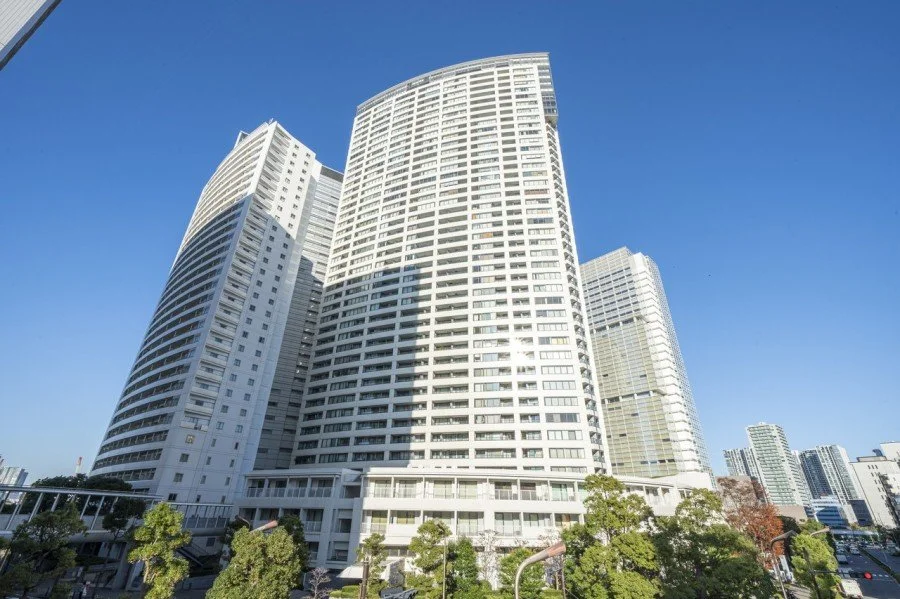 高層の白いビル群と青空、下に樹木と道路が見える都市の景観