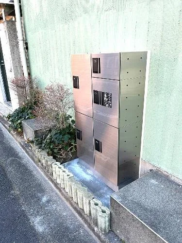 路側に設置されたコンクリート製の郵便ポストと宅配ボックスの複合式収納箱。背景は淡い緑色の壁と植物が見える。