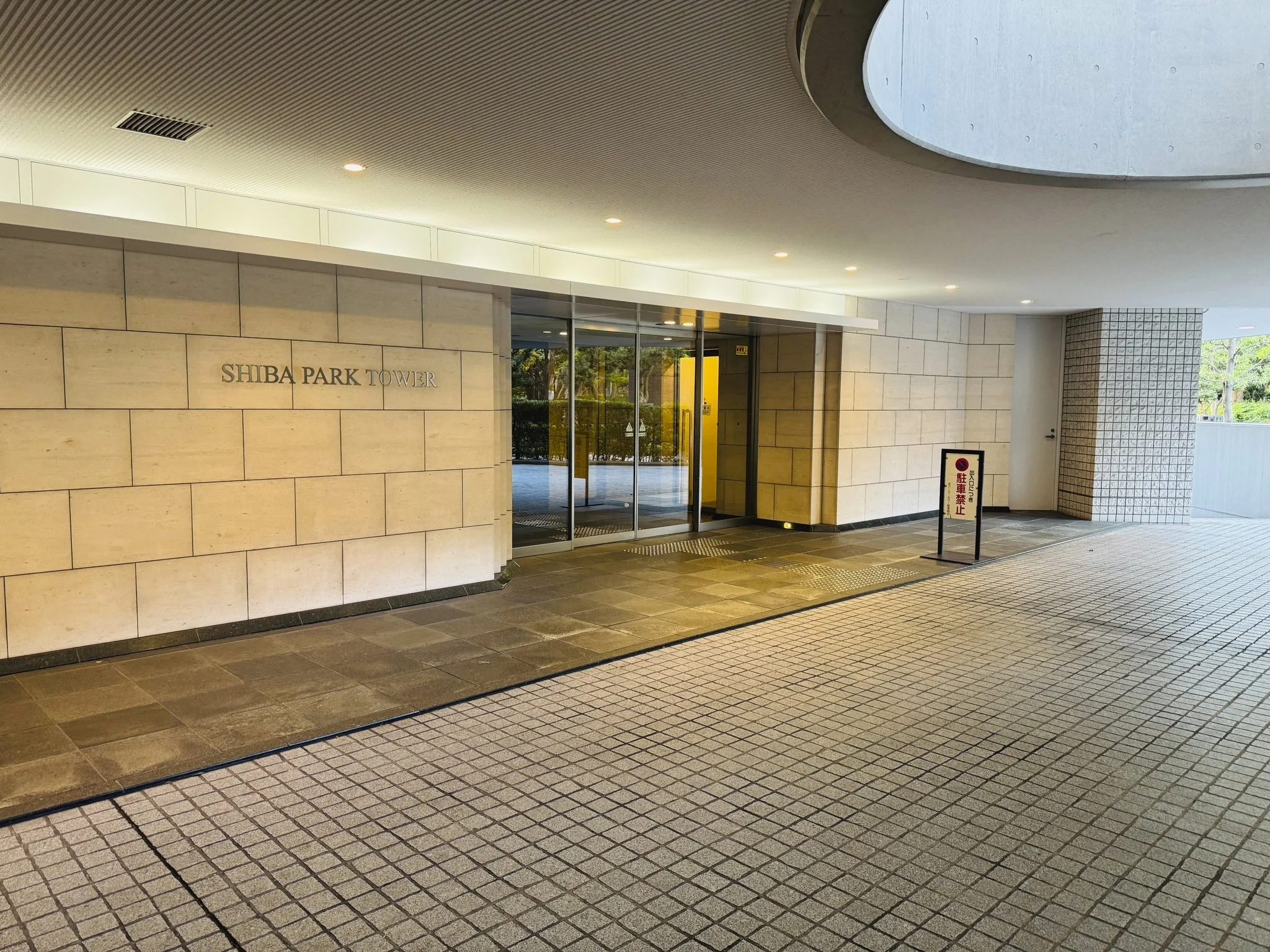 東京のシバパークタワーの入口。壁に「SHIBA PARK TOWER」のサインがあり、ガラスドアが見える。舗装された歩道と小さな立て看板も映っている。