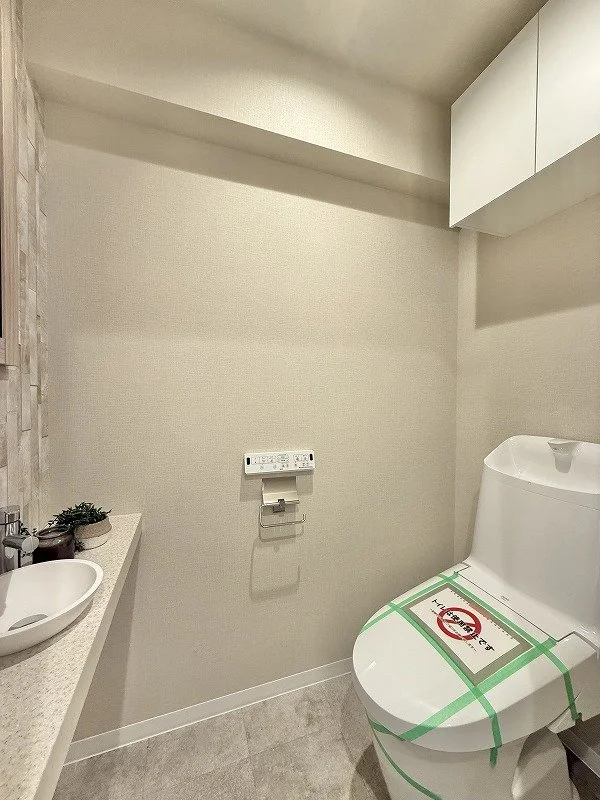 トイレと洗面台があるシンプルなバスルーム。便座には緑色のテープと注意書き付き。