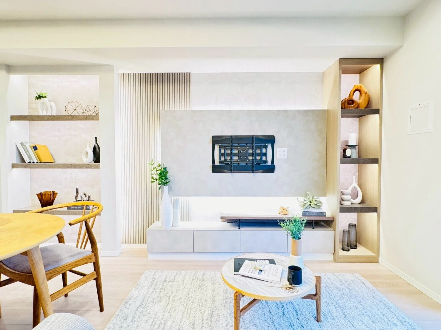 シンプルでモダンなリビングルーム。壁にはテレビ掛けが付いている。棚には装飾品や本が飾られている。コーヒーテーブルに花瓶とマグカップが置かれている。木製の椅子とダイニングテーブルも見える。部屋は明るく、ナチュラルな色調で統一されている。