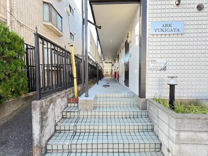 マンションの入口の階段と廊下。右側に青い表示板に「ARK YUKIGAYA」と書かれている。