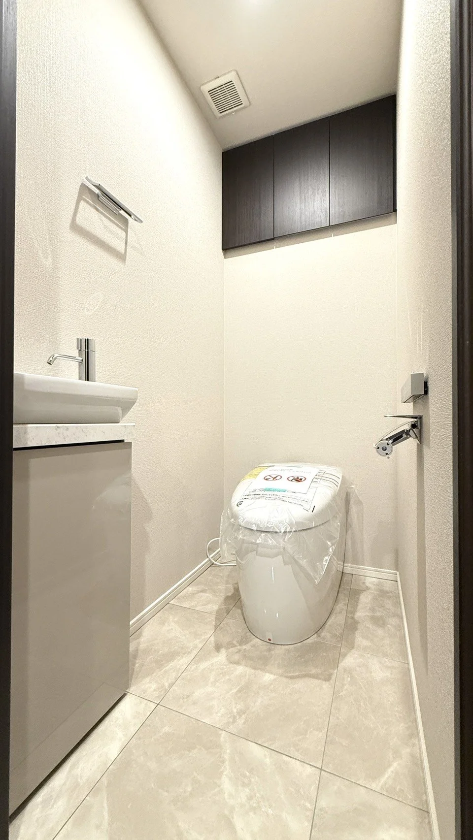 新しいトイレと洗面台があるシンプルなトイレ室内の写真。壁にダークブラウンの収納棚と、天井に換気扇が見える。