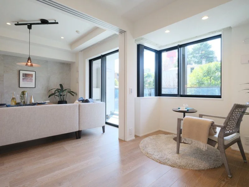 明るいリビングルームのインテリア。ソファ、ラグ、丸いテーブル、チェア、観葉植物、大きな窓とガラスドア、屋外の植栽が見える。