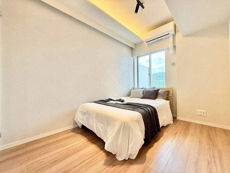 明るい照明のあるシンプルな寝室で、白いベッドに灰色と白の枕が置かれ、窓の外にビル群が見える部屋