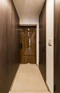 廊下の写真、左側は暗い木目調の壁、右側は白い壁と扉、遠くに照明のある木製のドアが見える。