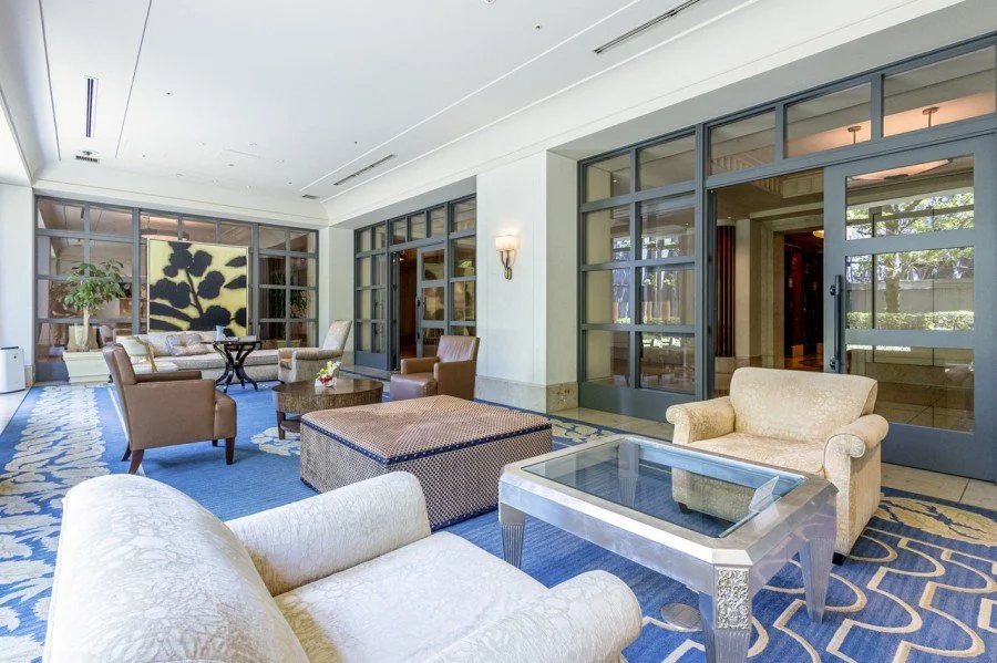 広々としたリビングルームにソファ、アームチェア、コーヒーテーブルが置かれ、ガラス扉の向こうに緑の木々と外の景色が見える。