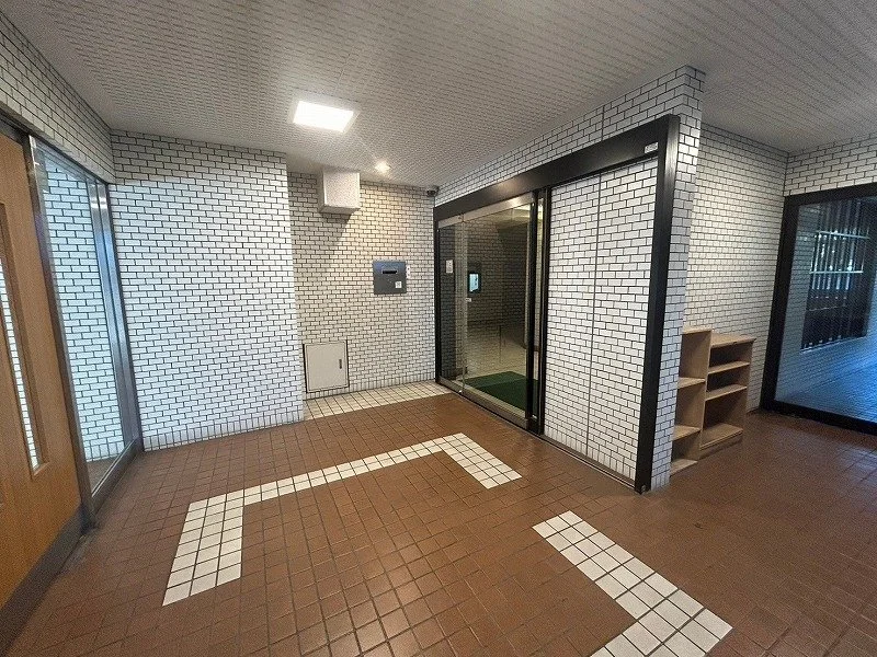 白いレンガの壁とドアのある建物の入口。床には白いラインと矢印のマークが描かれたタイル。右側に木製の棚が置かれている。