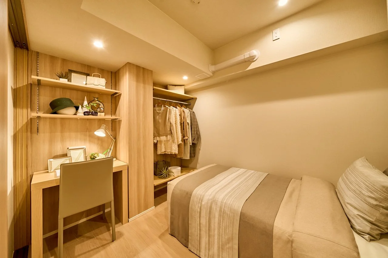 シンプルなデザインの小さな部屋、シングルベッドと木製の机、棚には帽子や写真立て、クローゼットには洋服と植物が掛かっている。