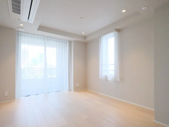 白い壁と木製のフローリングの空室の部屋。窓には白いカーテンが掛かっている。