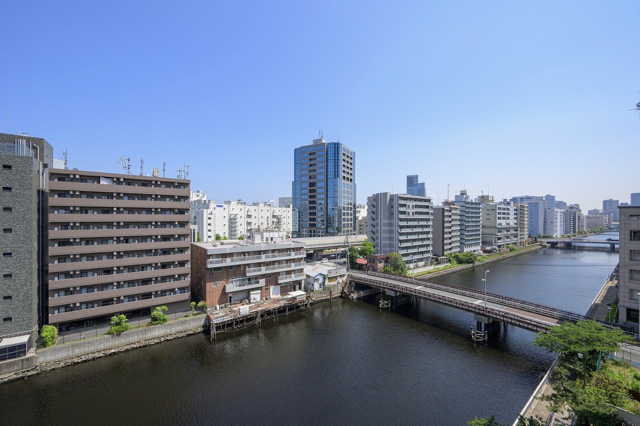 都市の川沿いに建つ多層ビルと橋、青空が広がる風景