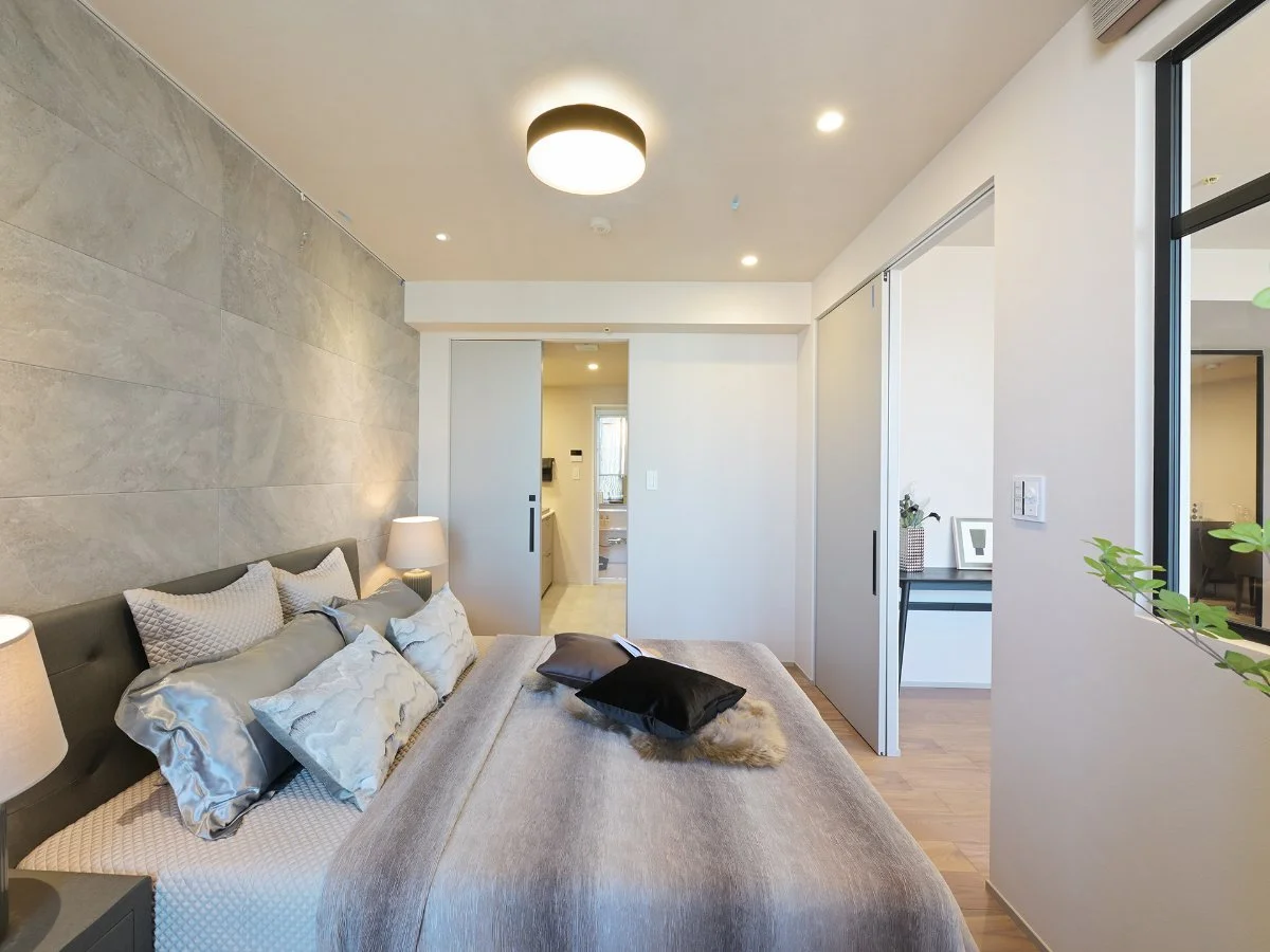 モダンな寝室の内側の写真。ベッドにクッションと毛布があり、壁には照明と窓があります。