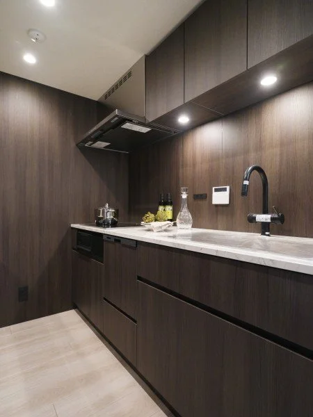 モダンなキッチンの内部、木目調のキャビネットと白いカウンター、シンク、蛇口といくつかの調味料ボトルが置かれている。