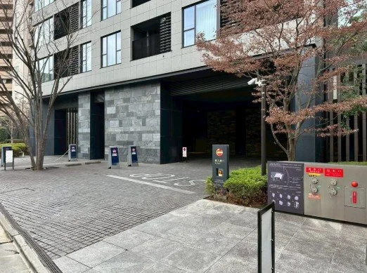 都市の建物の入り口付近の風景。舗装された通路とゲート、案内表示板や機械設備が見える。木の葉が紅葉している。