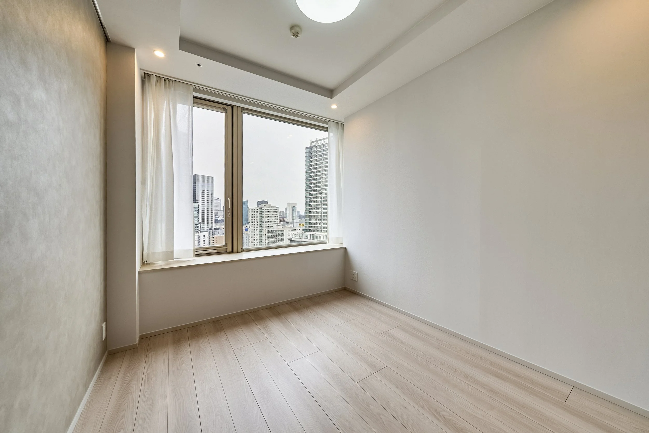 白い壁の部屋に大きな窓と市街地の高層ビルが見える風景。