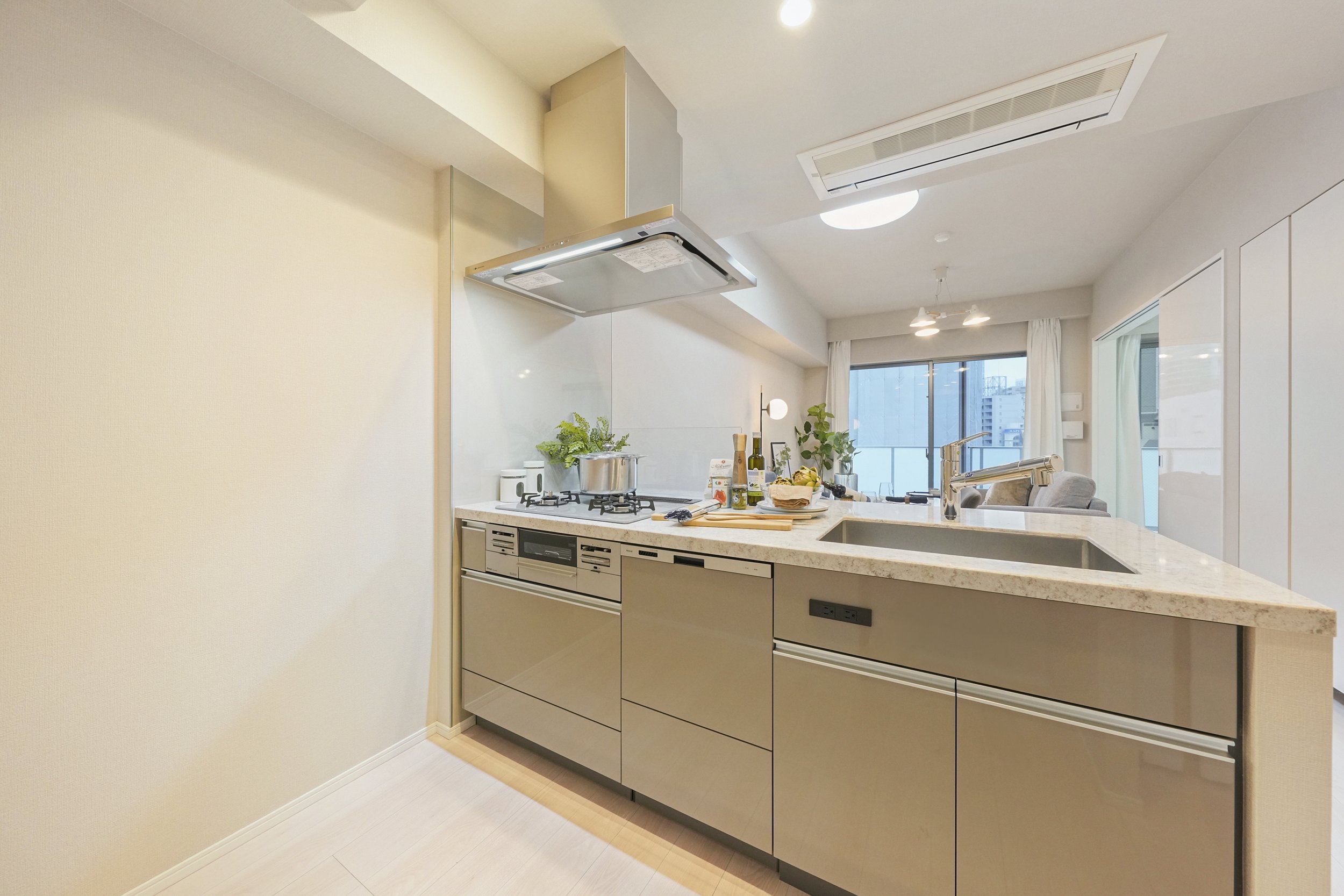 モダンなキッチンの interior with beige cabinets, a marble countertop, a stainless steel sink, and cooking area with a pot, plant, and kitchen utensils. background shows a living room with large windows and a balcony.