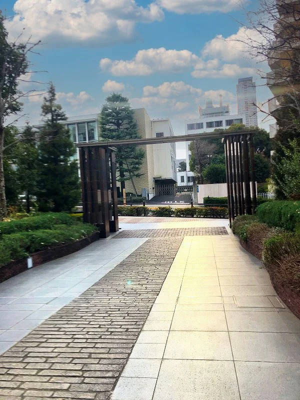 都会の建物が見える庭園の入り口で、木の門と緑の植栽があります。青空と白い雲も映っています。