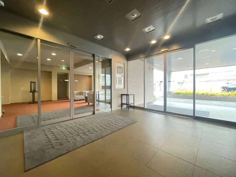 明るいロビーエントランス。ガラスの扉と窓の向こうには外の道路と車、室内には椅子と壁に掛けられた写真が見える。