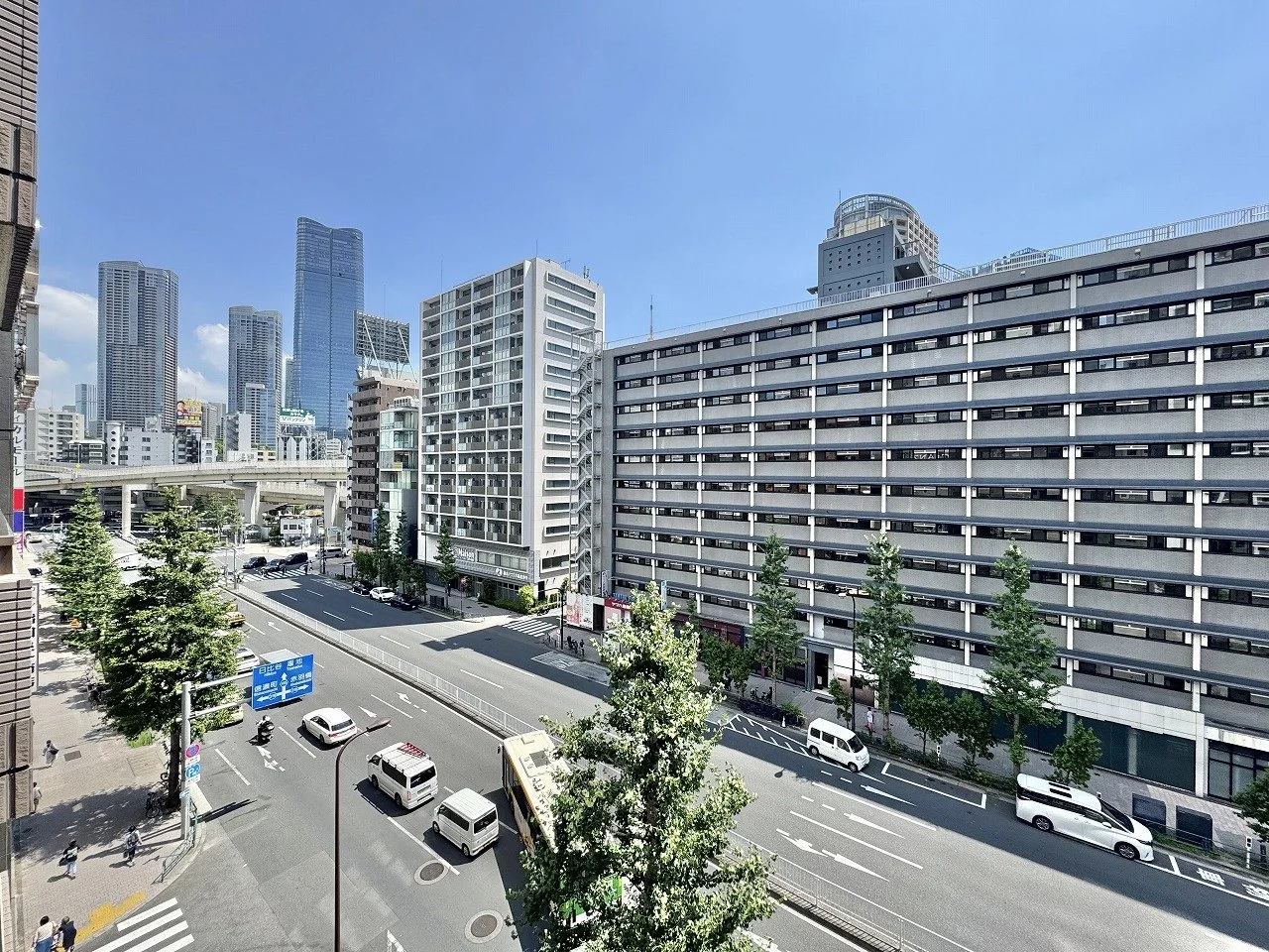 高層ビルが立ち並ぶ都市の道路風景。道路には車やバイクが走っており、歩行者が横断歩道を渡っている。周囲には樹木が植えられている。