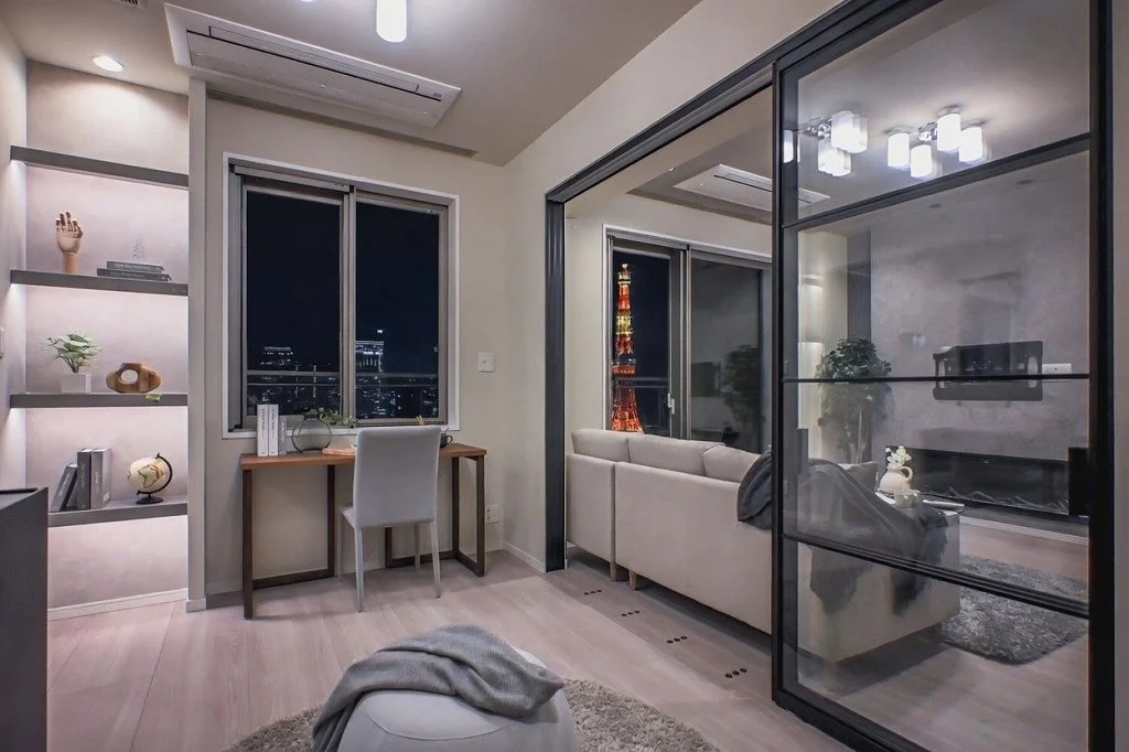 夜のリビングルーム。窓の外には東京タワーが見える。ソファ、テレビ、棚に装飾品や books あり、部屋はモダンなインテリアで明るく整っている。