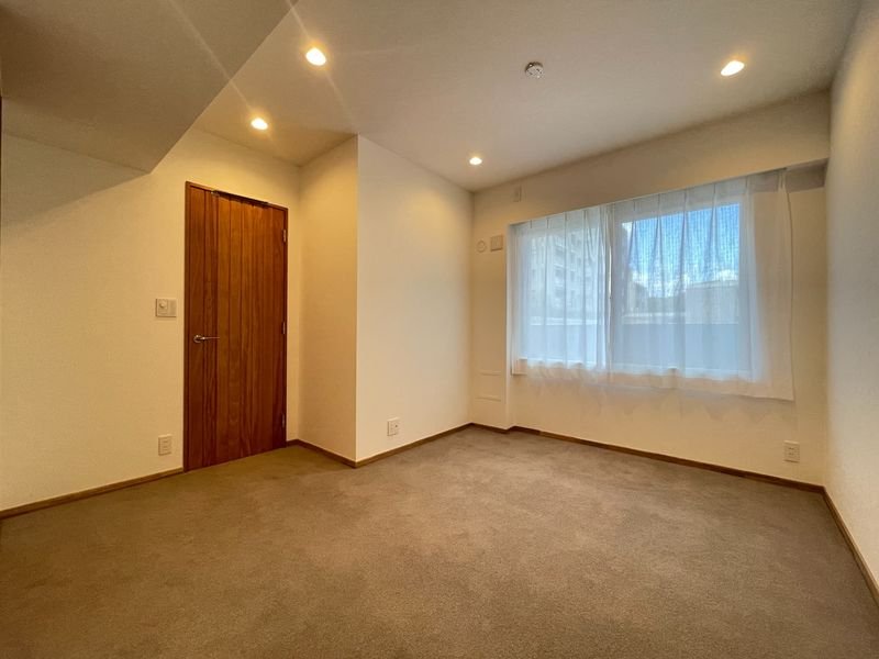 明るい照明のついた白い壁とカーペットの部屋、木製のドアと大きな窓、白いカーテンがかかっている