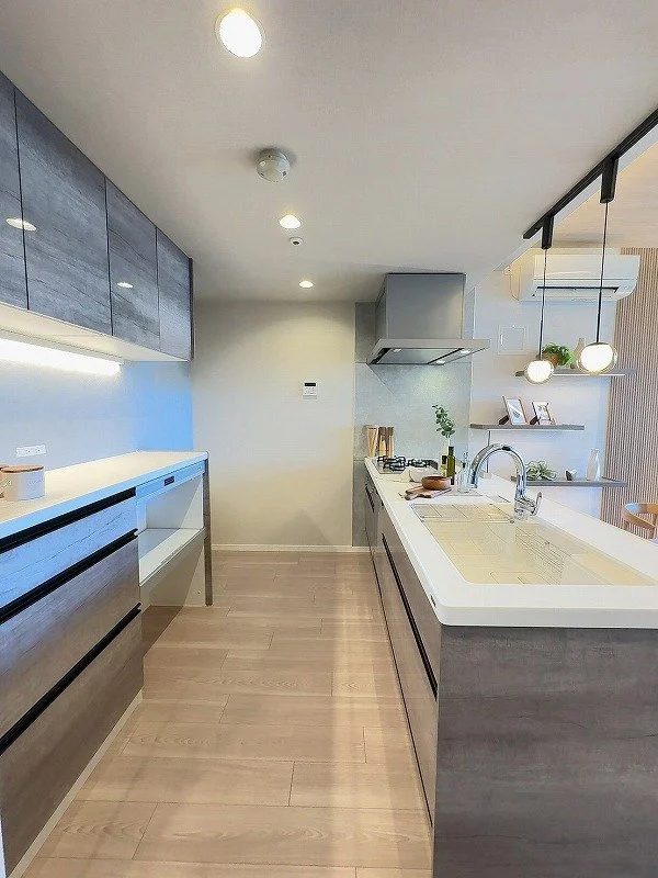 モダンなキッチン、白い作業台、木製の床と収納、壁に棚と植物、吊り灯篭。