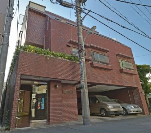 赤レンガ造りの三階建ての建物で、駐車場に車が2台停まっている。上部には電線も見える。