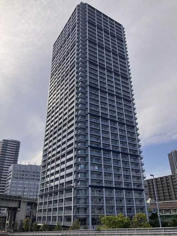 高層ビルの外観。ガラスと金属を使った現代的なデザインの建物。青空と数軒の低層建築物が背景に見える。
