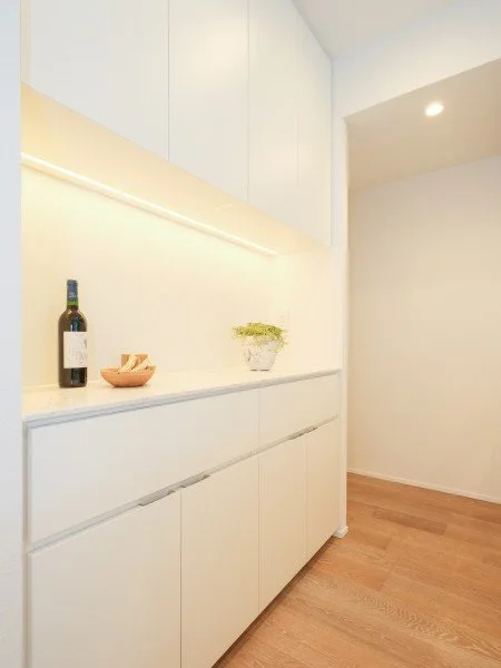 白いキャビネットの上にワインのボトルと小さな皿、植物が置かれているキッチンの一角。