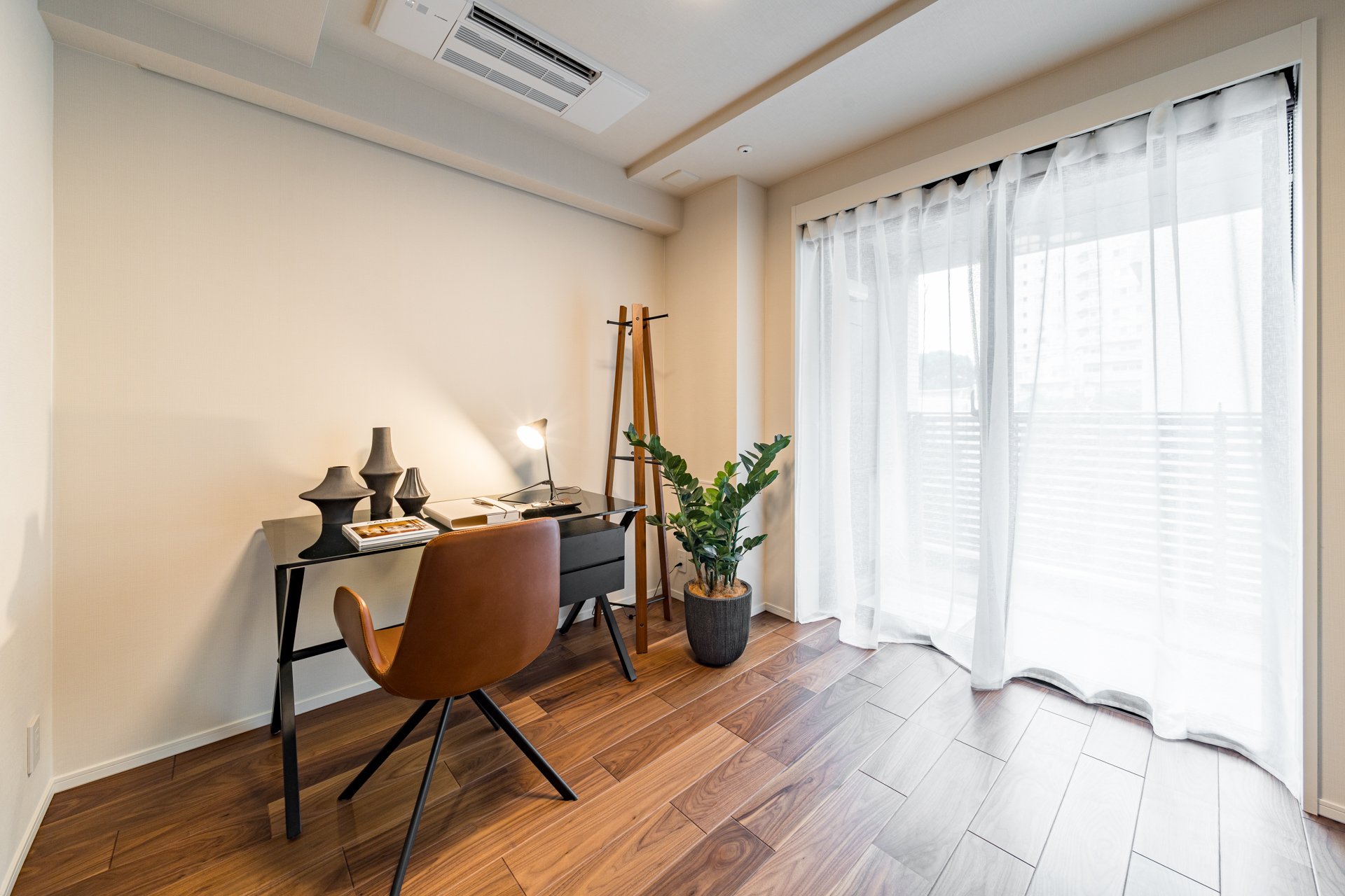 シンプルな書斎スペースに黒い机と茶色い椅子、観葉植物、白いカーテンの窓がある部屋の写真