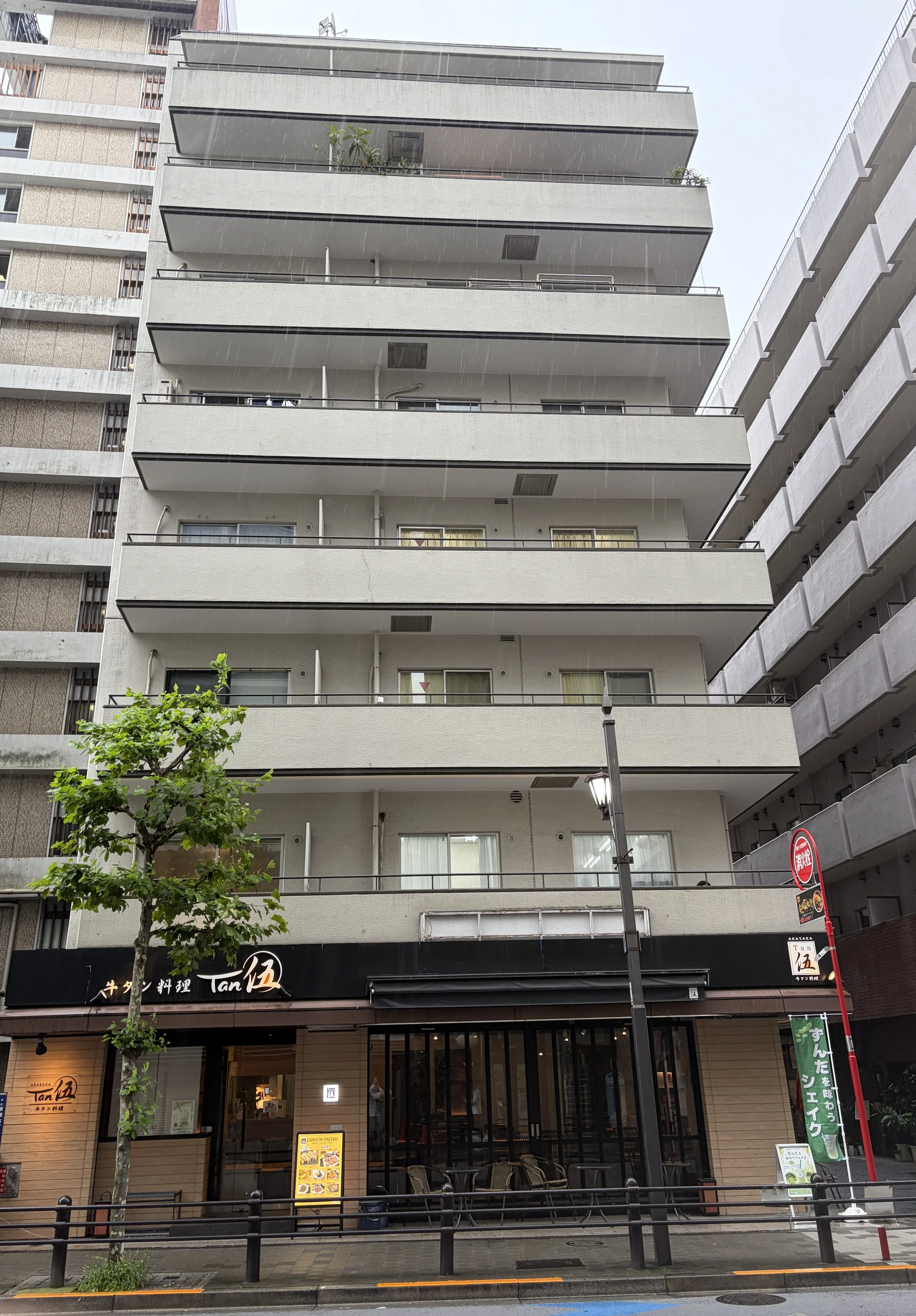 都市のビルの1階にレストラン店の看板と入り口、外にテーブルとイス、左側に木の木があり、雨が降っている。