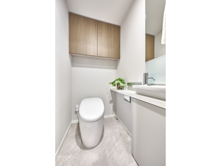 シンプルでモダンなトイレルーム、白い便器と洗面台、木製の収納棚と緑の観葉植物が配置されている