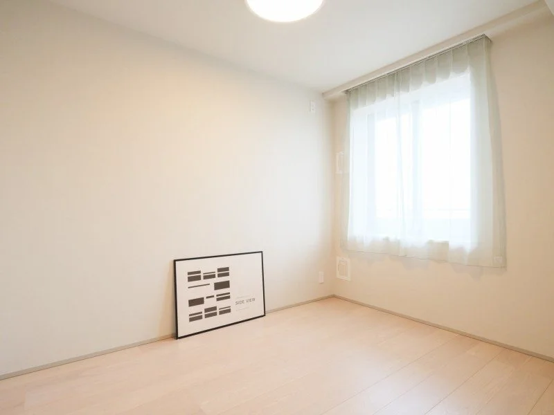 白い壁のシンプルな部屋で、窓には薄いカーテンがかかっている。床は明るい木製のフローリング。壁に黒いフレームのアートが立て掛けてある。