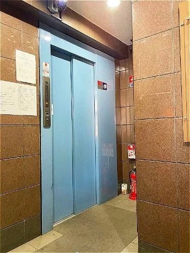 青色のエレベーターの扉と階数表示パネルが壁に取り付けられたエレベーター前の壁。左側に掲示物、右側に消火器があります。
