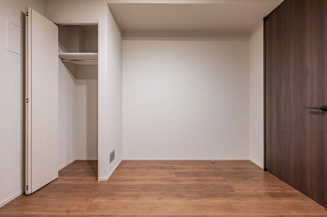 空の部屋、左側に白い折りたたみ式のクローゼットドア、床は木製のフローリング、右側にダークブラウンの木の扉
