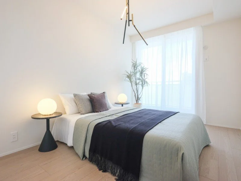 明るい寝室で、ベッドには白とグレーの寝具と紫のクッションが置かれている。サイドテーブルには丸い白いランプが二つあり、背後には大きな窓とカーテンがある。室内には観葉植物も置かれている。