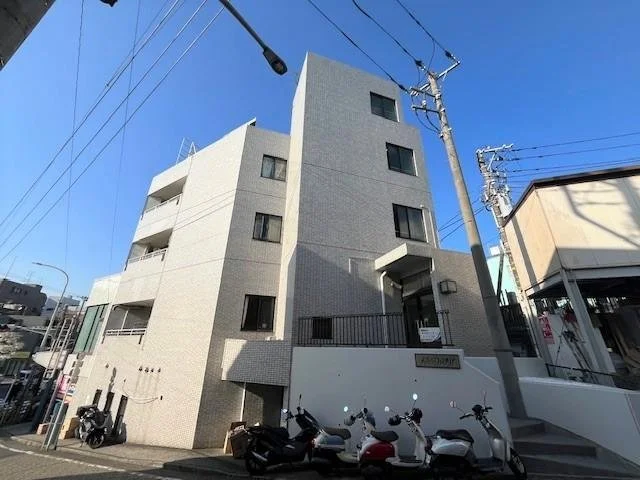 多層の灰色の建物と電柱、電線、複数のバイクや自転車が停められている道路。青空。