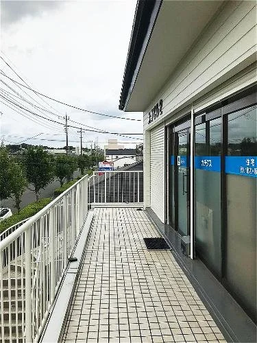 建物の屋上のテラスから見た風景。バルコニーには白い手すりとタイルの床があり、ガラス扉と窓が見える。外には電線と木々が見える。
