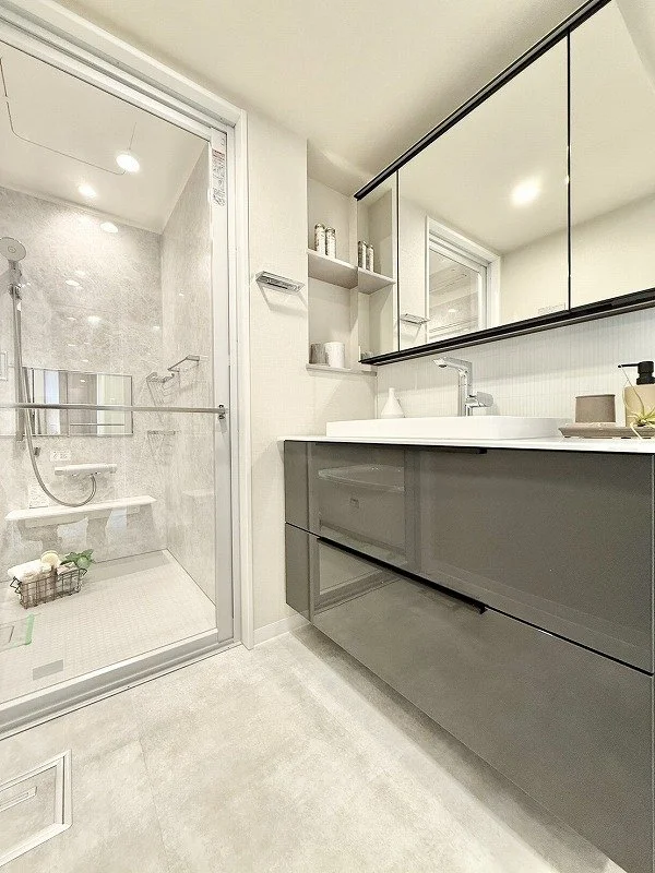 白と灰色のモダンなバスルームと洗面台の鏡張り鏡。シャワーエリアにはシャワーチェアとバス用品が配置されている。