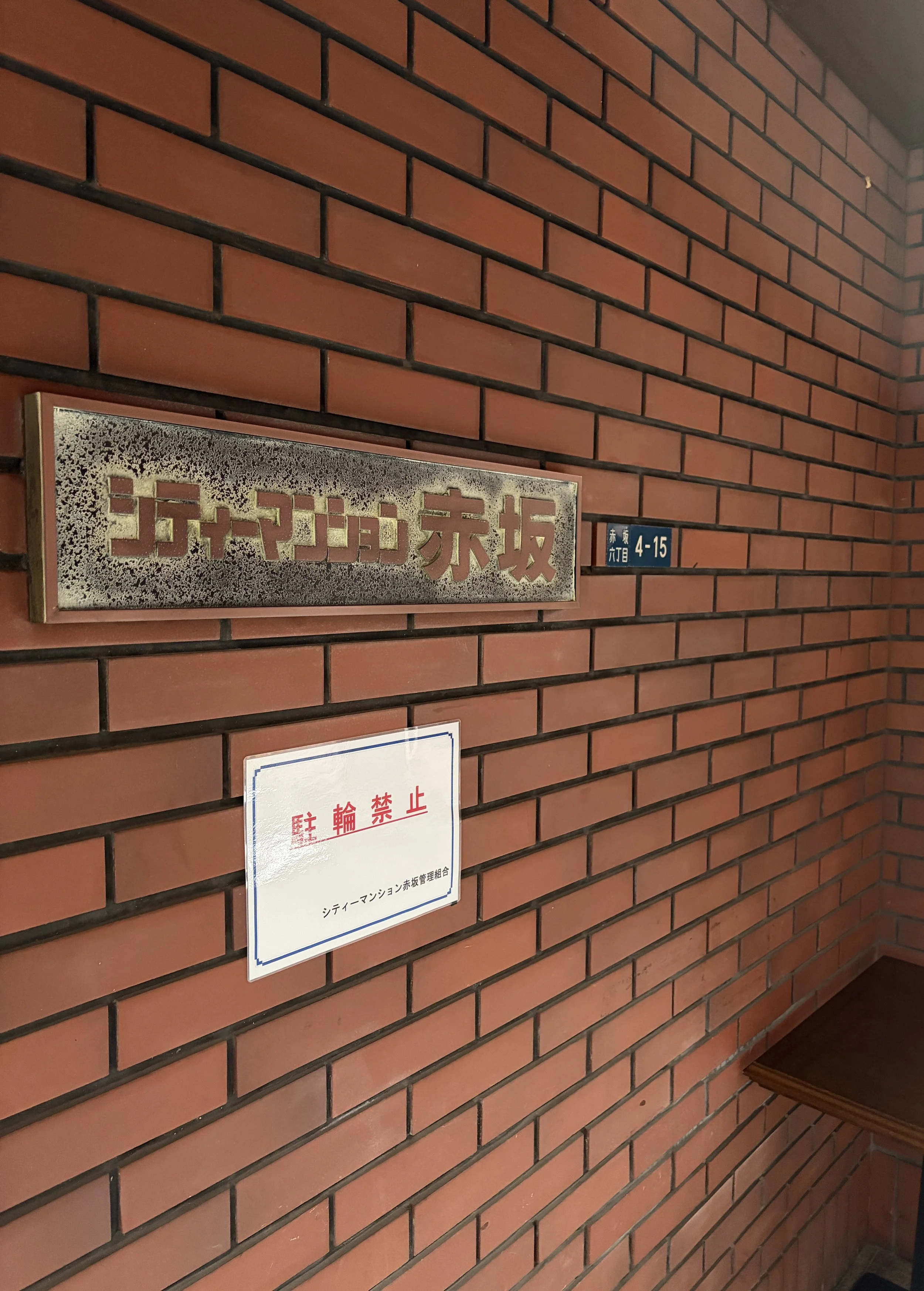 赤レンガの壁に掲示板と看板がある。掲示板には金属製のプレートに赤字で「赤坂元坂」と書かれている。横に青いプレートに住所のような文字列がある。