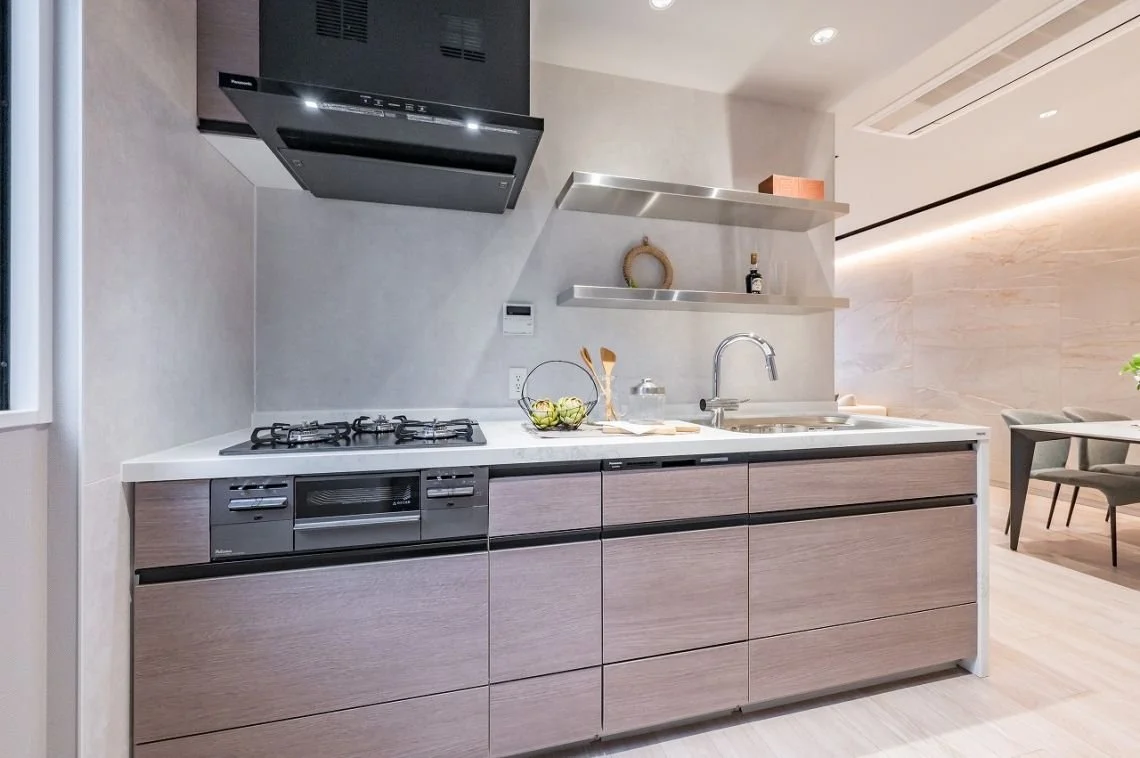 モダンなキッチンで、コンロとシンクがあり、棚に装飾品と調味料が置かれている。背景にはダイニングエリアが見える。