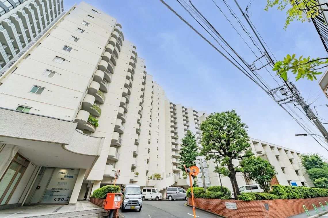 高層の白いマンションと木のある道路風景。青空と電線も写っている。