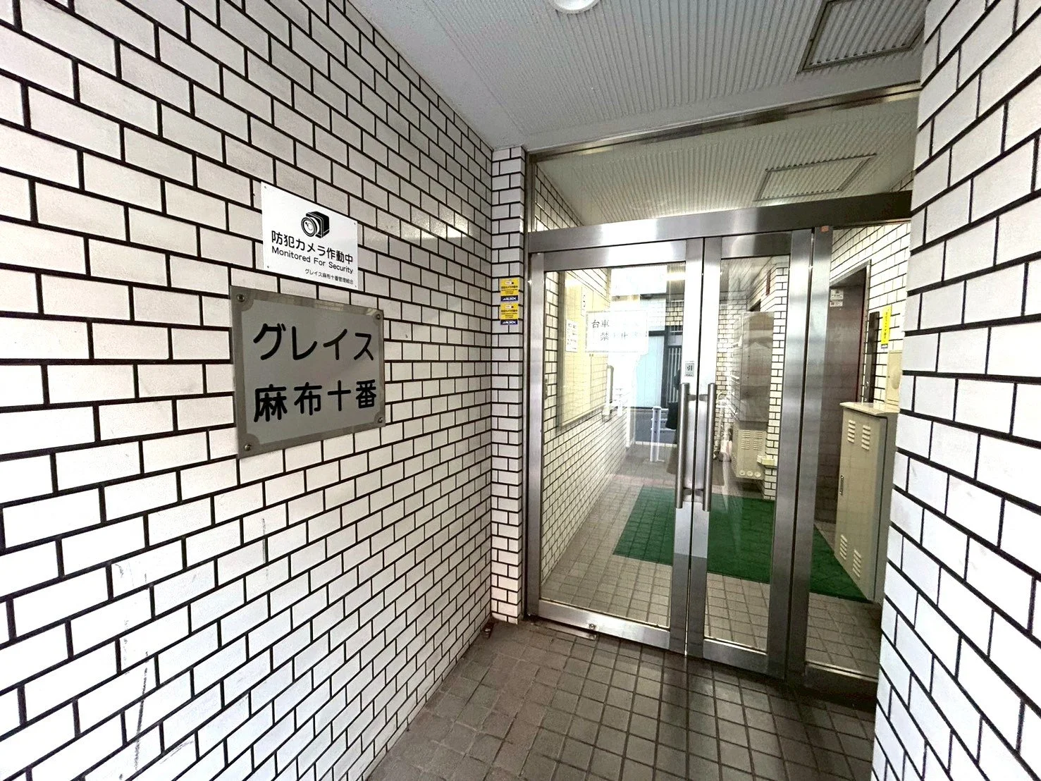 白いレンガの壁に「グレイス 麻布十番」と書かれた掲示板と防犯カメラのモニター表示がある入口ドアの入り口