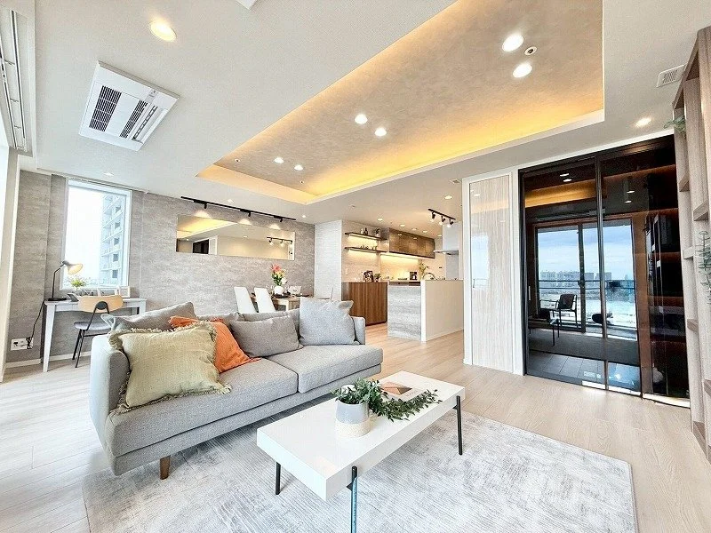 明るく広々とした現代的なリビングルームで、ソファ、コーヒーテーブル、ダイニングセット、シェルフ、バルコニーのガラス扉が見える。