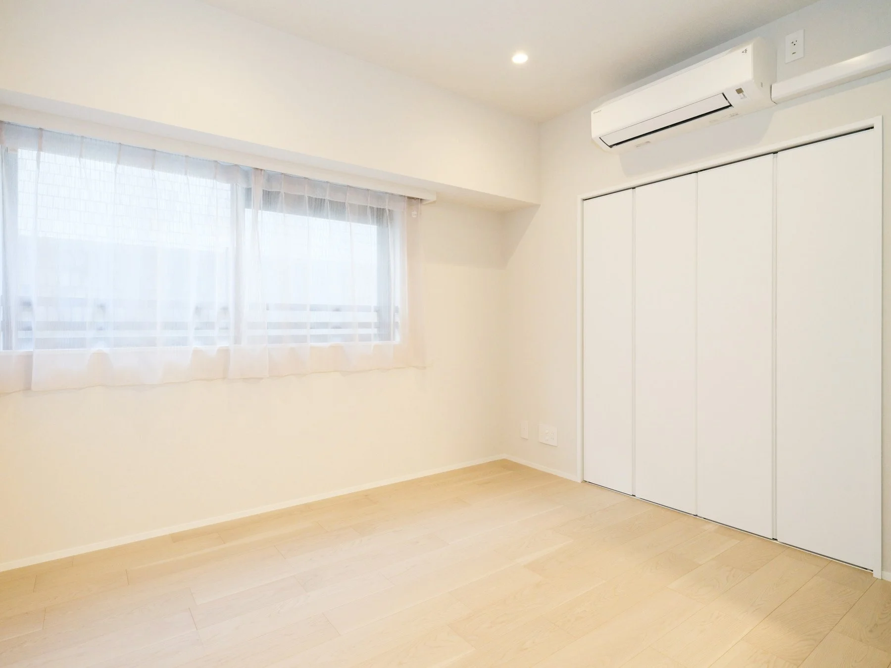 白い壁と木製のフローリングの部屋。窓には白い薄いカーテンが掛かっている。クローゼットの扉は白色。天井にはスポットライトとエアコンが設置されている。