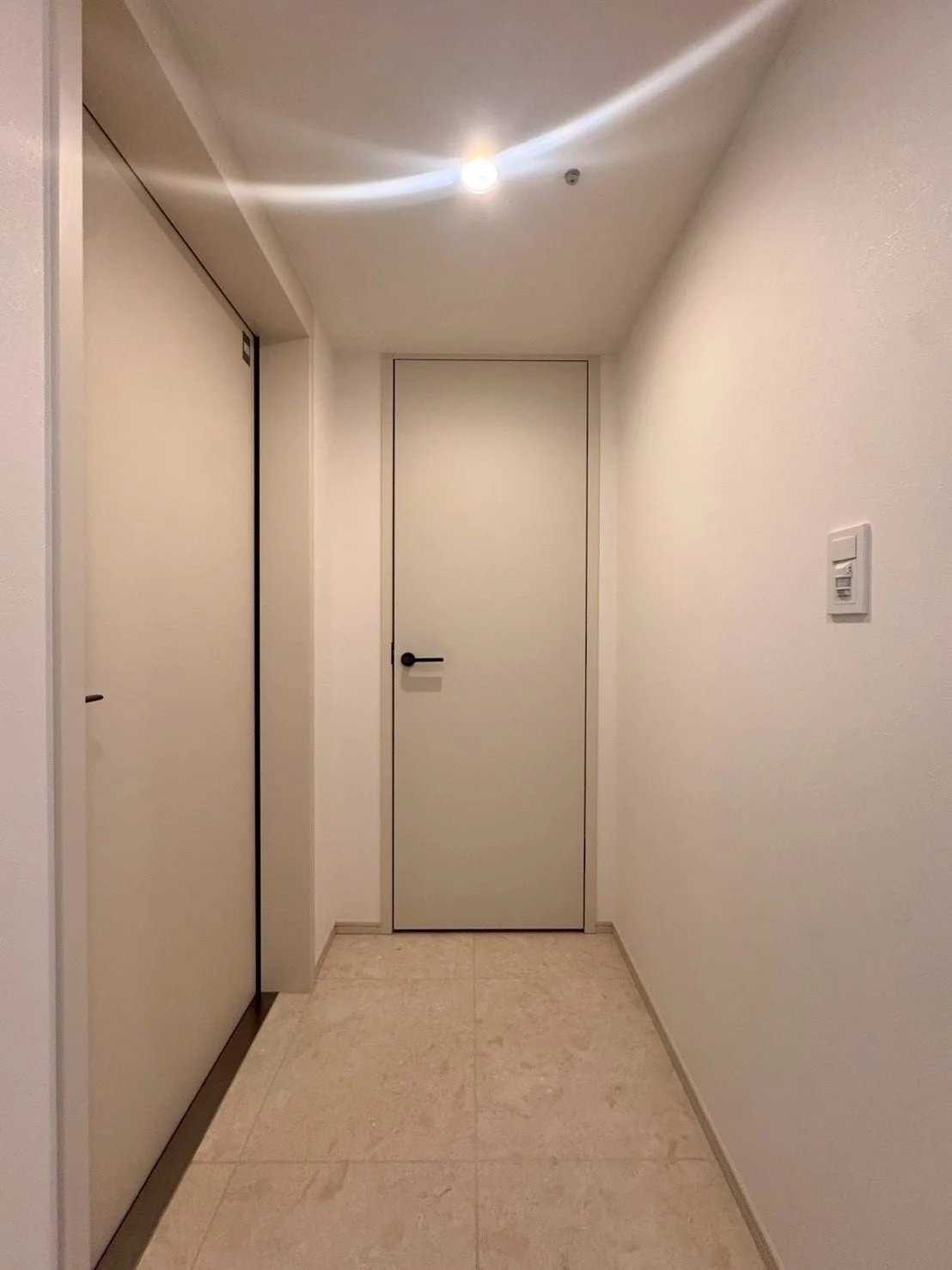白い壁と扉がある廊下の画像。左側は二つのドア、中央は閉じられたドア、右側にはスイッチがある。