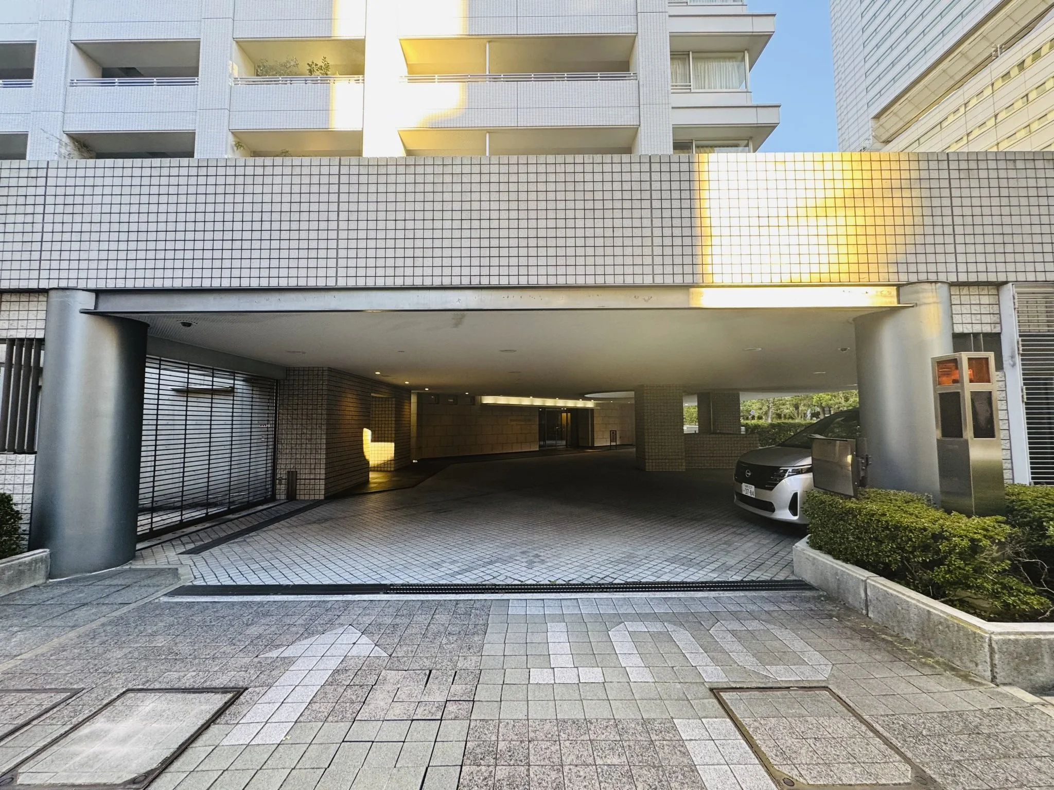 コンクリート造りの駐車場の入り口と駐車スペース。左側には車止めとやや閉ざされたシャッターあり。右側には駐車中の白い車と自動販売機。背景には多層階の白いマンション。