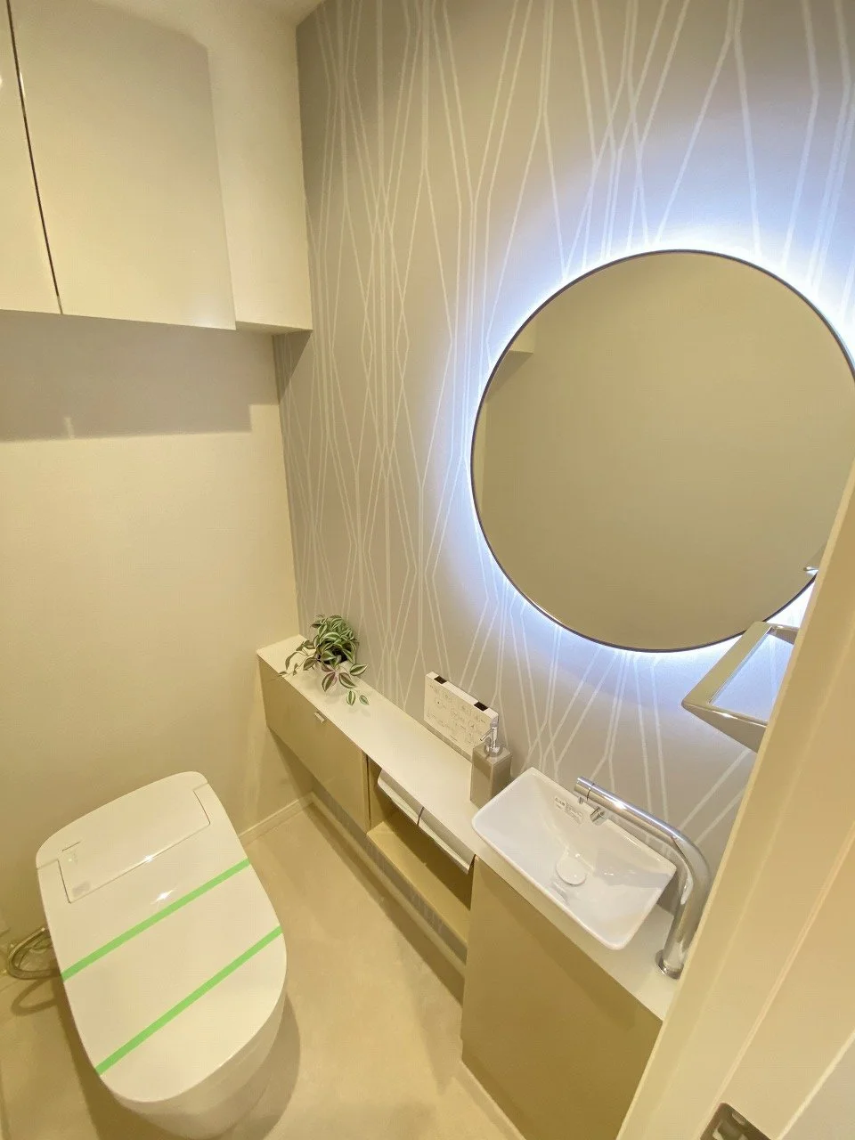 シンプルなモダンなトイレルーム。丸い鏡と洗面台、植物と収納棚があります。