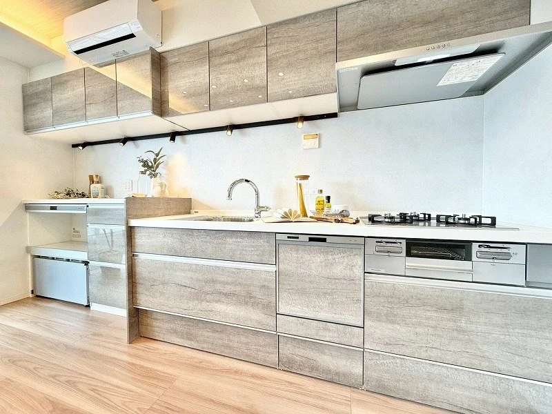 モダンなキッチンの内部、木目調のキャビネットと白いカウンター、ガスコンロとシンク付き、掃除用品や調味料が置かれている