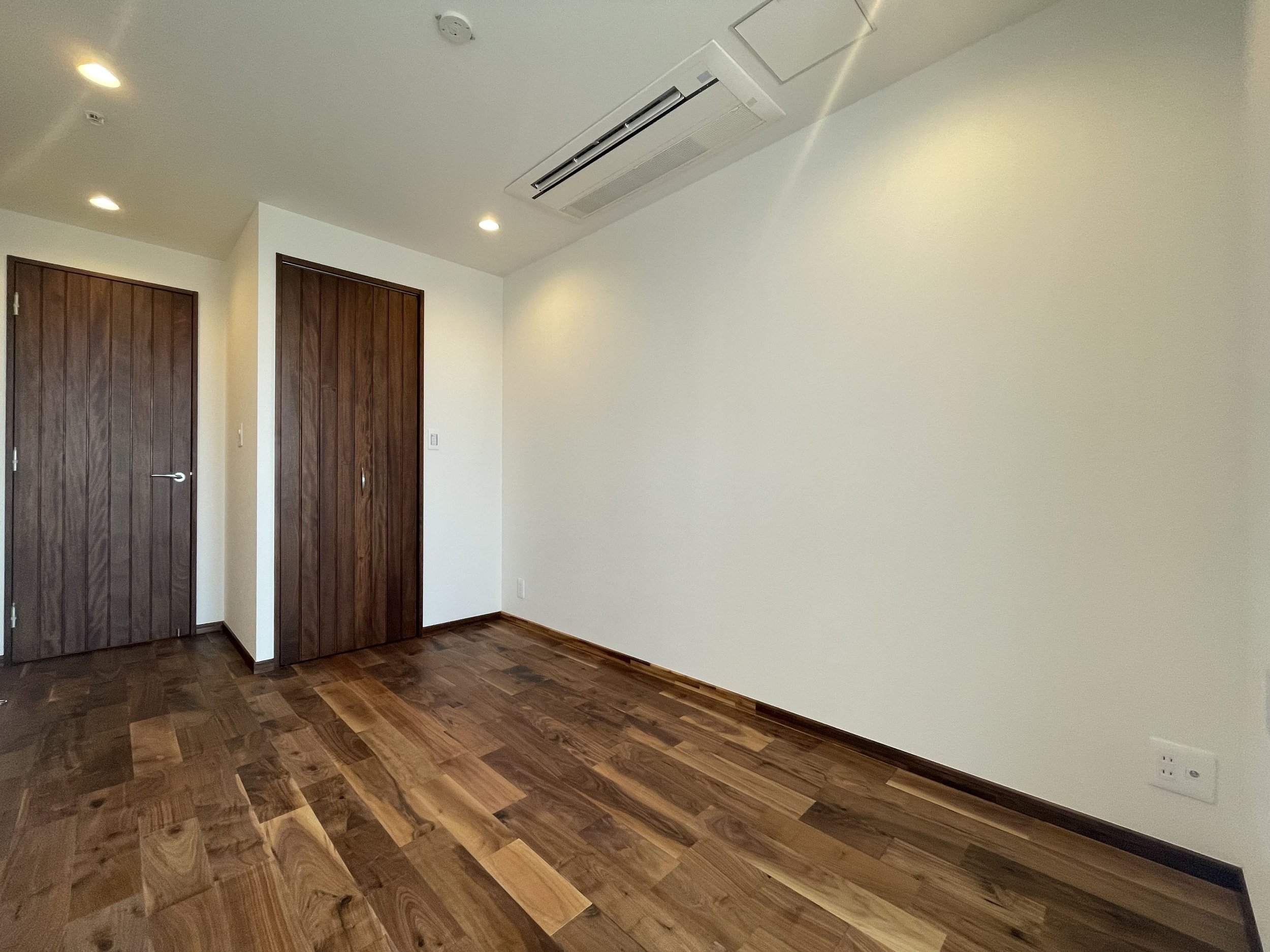 木製の床と白い壁の空き部屋。ドアとクローゼット扉は木製。電気コンセントとエアコンが設置されている。
